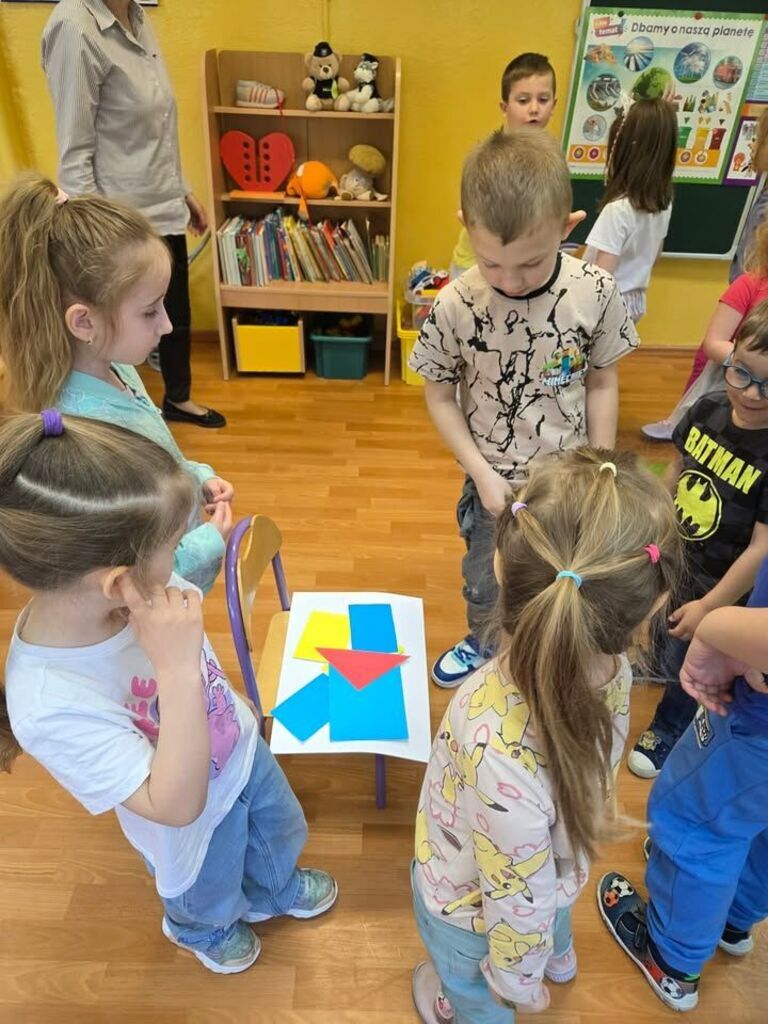 dzieci stoją wokół krzesła na którym leża wycięte z kolorowych papierów figury geometryczne