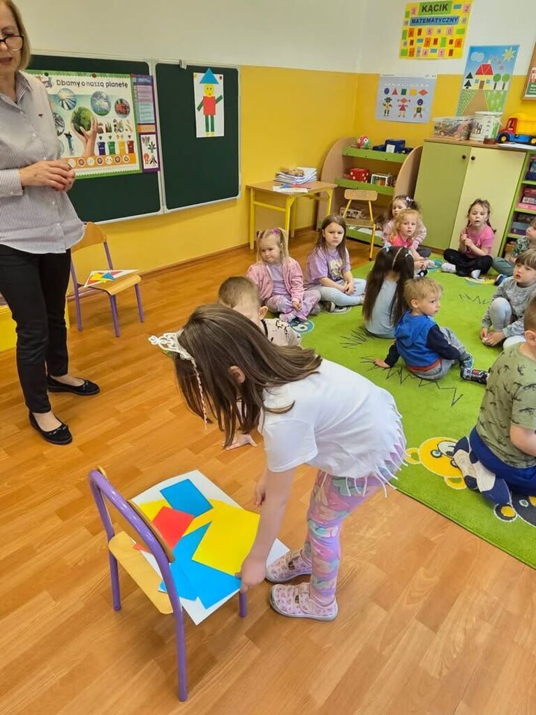 dzieci siedzą w kółku na dywanie, dziewczyna wybiera kształt geometryczny wycięty z kolorowego papieru